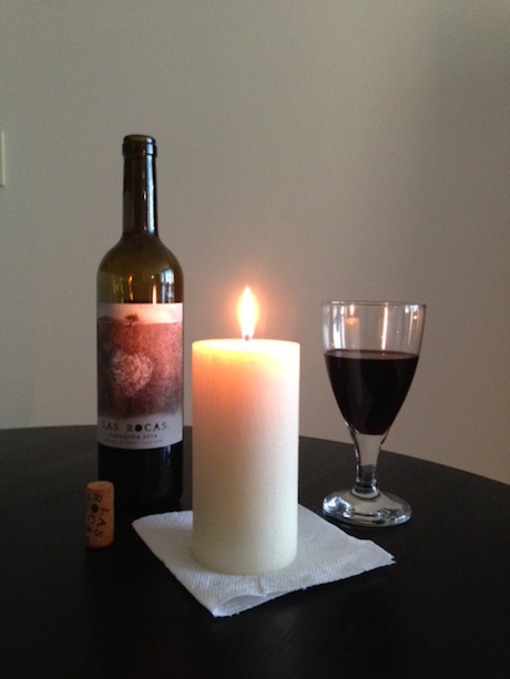 A photographic image of an opened bottle of Las Rocas red wine together with a lit candle and water goblet filled half-way with the wine.